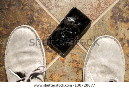 A smartphone lies broken between the shoes of its owner just after being dropped.