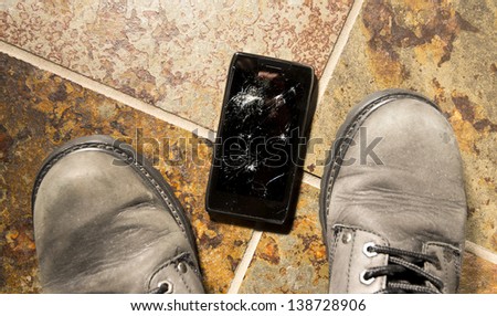 A smartphone lies broken between the workboots of its owner just after being dropped.