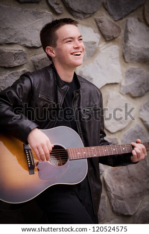 Teenage Caucasian boy with brown hair and brown eyes smiling and playing the guitar next to a rock wall