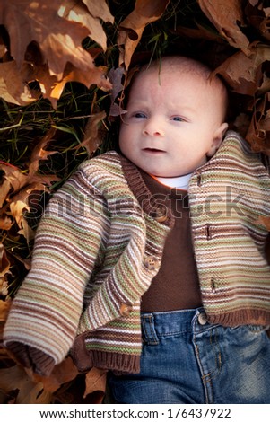 Curious newborn baby boy surrounded by fall leaves in a striped sweater and blue jeans