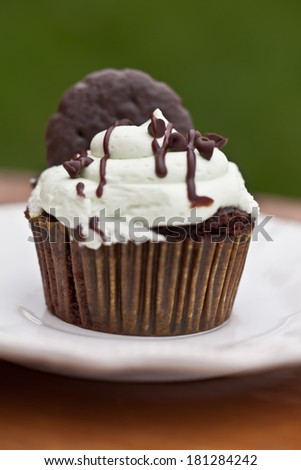 Delicious chocolate mint cupcake with miniature chocolate chips sprinkled on top topped with a chocolate mint cookie on a wooden table with a green grass background