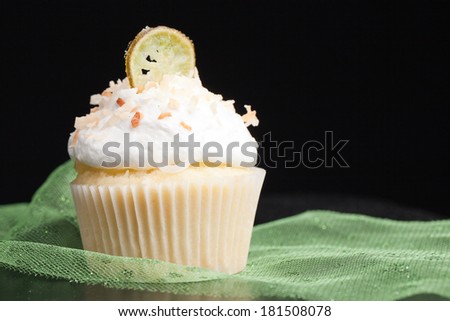 Coconut Key Lime Cupcake on a black background with green tule - Coconut cake with a key lime filling topped with a light 7 minute frosting, toasted coconut, and a slice of candied key lime