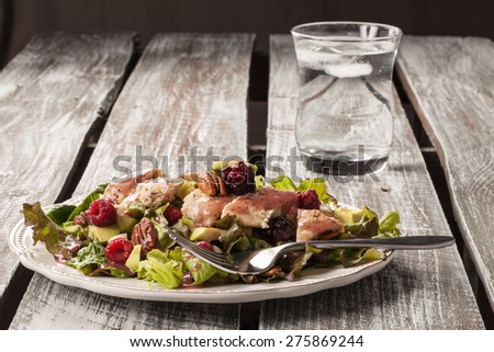 Side view of freshly harvested lettuce, grilled chicken, avocado, raspberry, pecan salad with a cup of water on an old weathered barn wood table