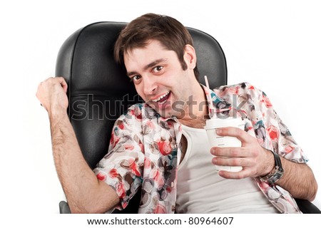A young handsome white guy sitting on a Caucasian black chair in a colorful Hawaiian shirt with a clock in his hand and holding a glass of drink in isolation over white