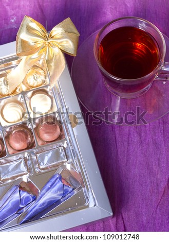 A box of chocolates with a bow around a cup of tea on a violet background