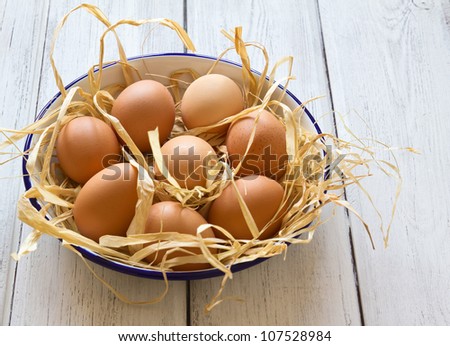 Eggs in enamel bowl on white wood background Eight fresh chickensÃ¢Â?Â? eggs on raffia in a white, blue-rimmed enamel bowl on a white-painted wooden background.