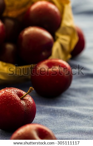Fresh Black Diamond plums in a brown paper bag on blue cloth background. Grown in Portugal. Whole fruits, one with a stalk. Some spilled from bag. Selective focus. Back lit. Studio shot, England, UK.