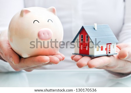 Hands Holding A Piggy Bank And A House Model. Housing Industry Mortgage Plan And Residential Tax Saving Strategy
