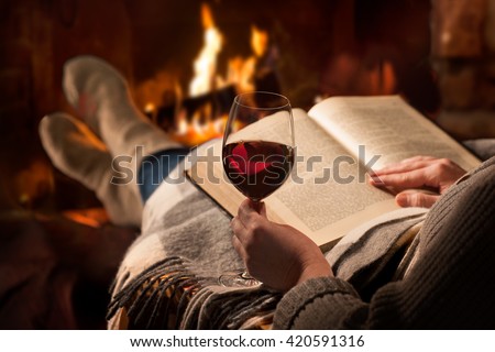 Woman resting with glass of red wine and book near fireplace