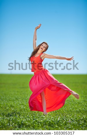 Beautiful young happy woman in long red dress dreams in green field against blue sky