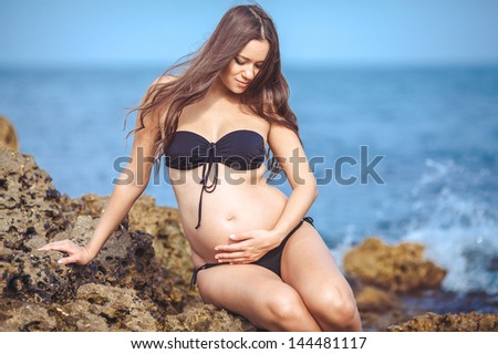 Beautiful pregnant woman outdoor in Greek goddess style with her tummy on sunset near sea. Young happy girl showing her belly and waiting for little baby. Pregnancy concept. Gorgeous pregnant lady