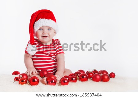 Adorable child is sitting on floor, wearing red Christmas cap, red balls around. isolated on white background