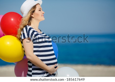 Portrait of beautiful pregnant woman in sunglasses and white hat with bright colorful balloons on sea shore background. Close-up