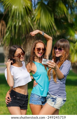 Group Of Four Teenage Girls Taking Picture In summer Park taking selfie photo using smartphone. Portrait of group of peolpe having fun outddors in green summer tropical park