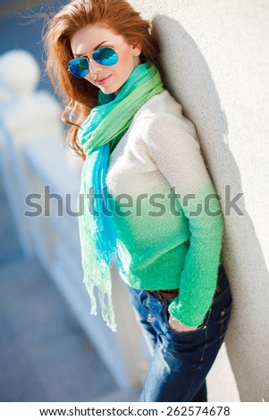 Beautiful modern woman with long red hair in a green-blue sweater and scarf outdoors. Woman with blue sunglasses. Smiling girl
