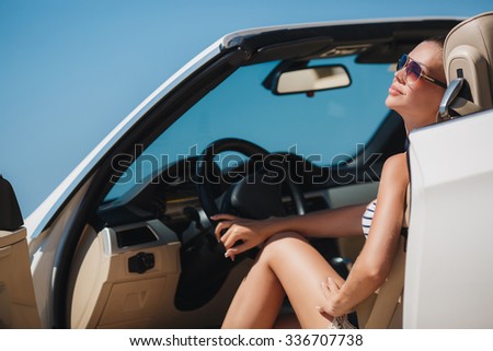 Portrait of beautiful sexy fashion woman model in sunglasses sitting in luxury white car with sea and sky background. Young woman driving on road trip on sunny summer day. Sea and sky. White cabrio.