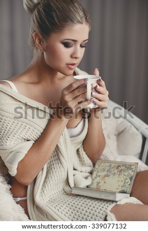 Young woman at home sitting on modern chair in front of window relaxing in her living room reading book and drinking coffee or tea. Autumn. Home. Cozy.Soft cozy photo of woman cup of tea in hands