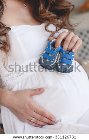 Small shoes for the unborn baby in the belly of pregnant woman. sneakers on belly. Pregnant woman holding small baby shoes relaxing at home.
