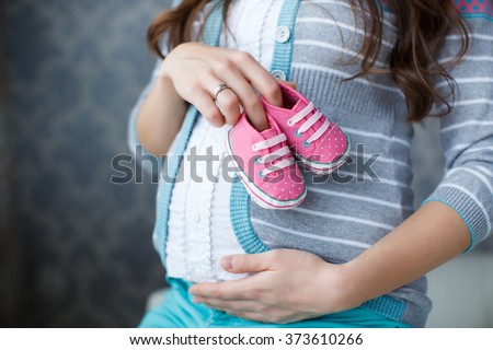 Small shoes for the unborn baby in the belly of pregnant woman. Pregnant woman holding small baby shoes relaxing at home in bedroom. Small shoes for the unborn baby in the belly of pregnant woman