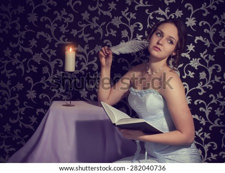Attractive romantic girl reading book and writing a poem and verses by feather (pen) by candle light as symbol of beauty, creation, art, education, studying, skill, seduction, sex, youth, adolescence