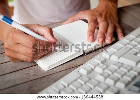Photo of hands writes a pen in a notebook, computer keyboard on foreground