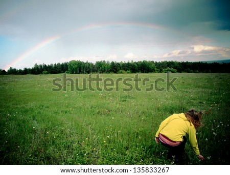 Young romantic woman from behind in a summer field picking flowers opposite to rainbow at horizon