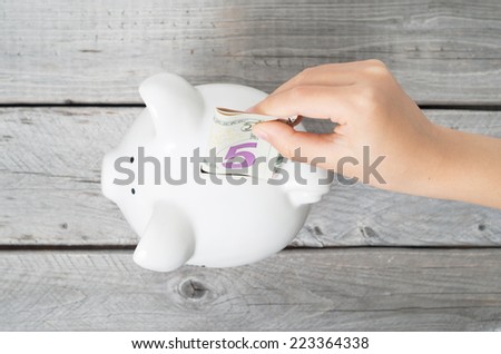 Asian boy hand inserting a five dollars bank note into white piggy bank  against wooden grey background
