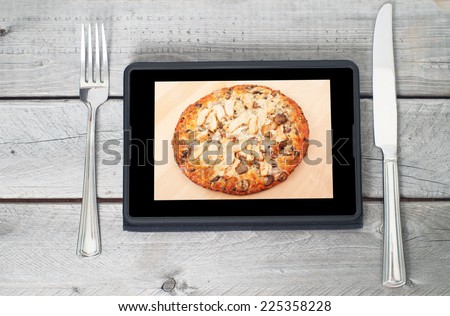 Tablet with an appetizing chicken pizza on its display between a fork and knife