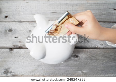 Asian boy hand inserting a hundred dollars bank note into white piggy bank  against wooden grey background