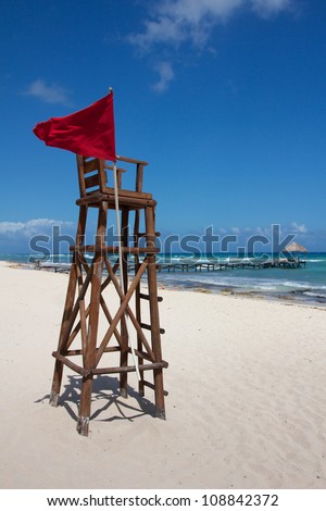 Empty lifeguard post with a red flag at a white, Caribbean beach under a clear sky.