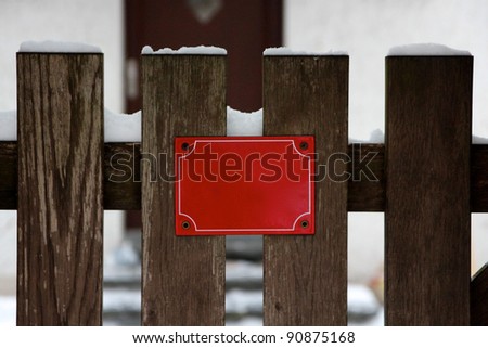 A small red sign mounted on a fence or gate of wooden planks. There is snow and a door visible in the background. The empty sign offers copy space.