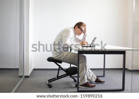 Young Man Is Bent Over His Tablet In His Office,Seating On Kneeling Chair Bad Sitting Posture At Work