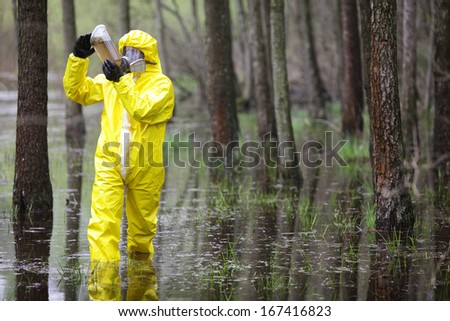 Fully Protected In Uniform,Boots,Gloves And Mask Technician Examining Sample Of Water In Plastic Container In Floods Area