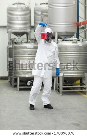 technician in protective uniform, mask, goggles, gloves examining liquid in plastic container
