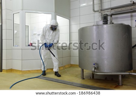 worker in white protective uniform,mask,gloves with high pressure washer at large industrial process tank cleaning floor in plant
