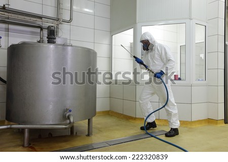 technician in white protective uniform,mask,gloves with high pressure washer at large industrial process tank cleaning floor in plant