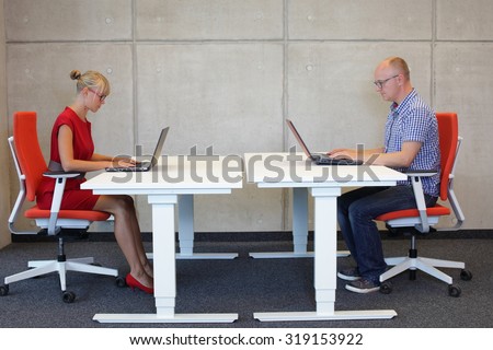 middle-aged man and young woman working in correct sitting posture with laptops at electric height adjustable desks in office