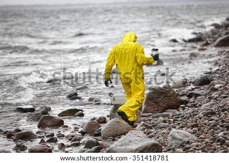 scientist in protective suit with plastic container walking in on rocky beach - back view