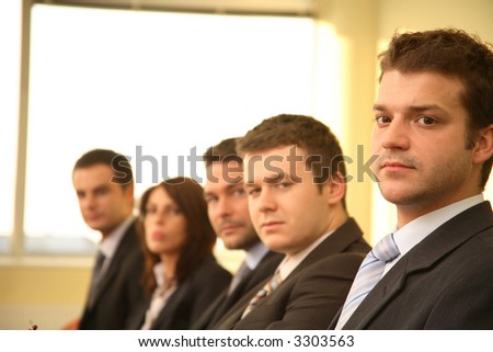 Five business persons in suits sitting at a conference table, taking part in a meeting and/or presentation.