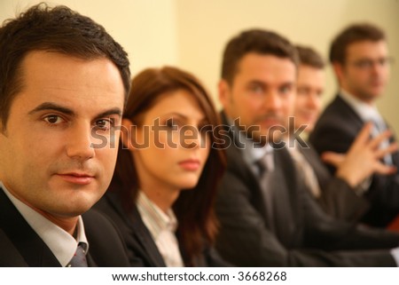 Group of business persons in suits sitting at a conference table, taking part in a meeting and/or presentation - one man in focus, portrait