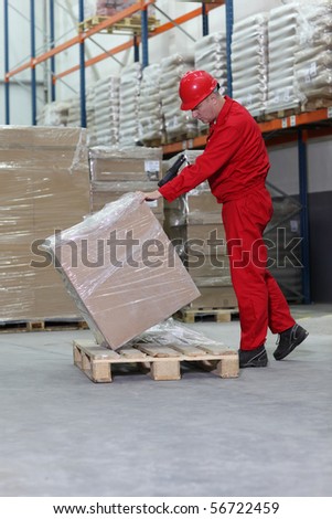 Middle aged workman in hard hat lifting cellophane wrapped box on wooden pallet in warehouse, stacked goods in background.