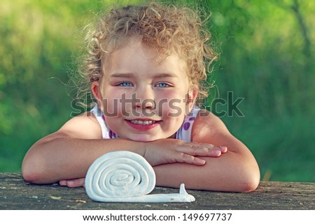 Little blonde girl rests her chin on her arms on the table and sees a snail on clay / The fast snail