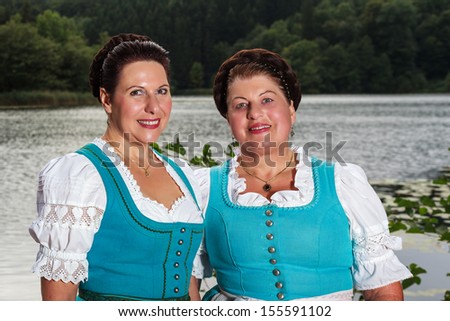 Two happy Bavarian ladies in dirndls standing side by side in front of a lake outdoors, upper body portrait / Two happy Bavarian ladies in dirndls