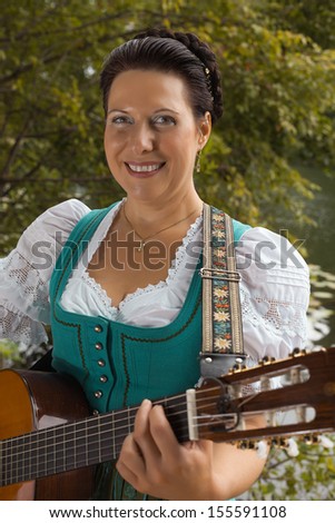 Smiling middle-aged woman in Bavarian Dirndl and braided her hair stands on the lake and playing guitar / Bavarian woman in dirndl smiling while playing guitar at the lake