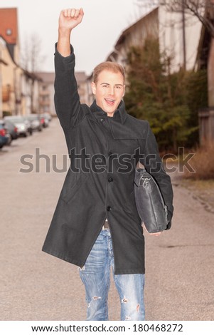 Handsome stylish successful urban man cheering as he walks down a street in town with a brief case under his arm and his arm raised in jubilation / Handsome successful urban man cheering