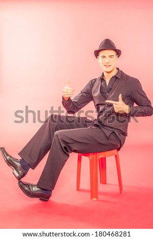 Handsome young man wearing a trendy hat floating mid air in the sitting position with his legs extended, conceptual image isolated on red / Handsome young man wearing a fashionable hat