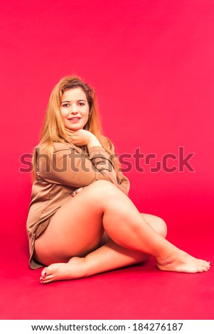 Obese beautiful young blond woman with long beige blouse and bare legs, sitting barefoot, smiling on the floor with a red background / Sensual image of a smiling young woman with overweight