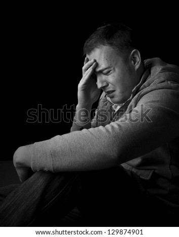man feeling pain, sitting on floor frowning with hand on head with black background