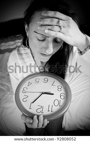 tired woman with clock waking late holding head sitting on bed