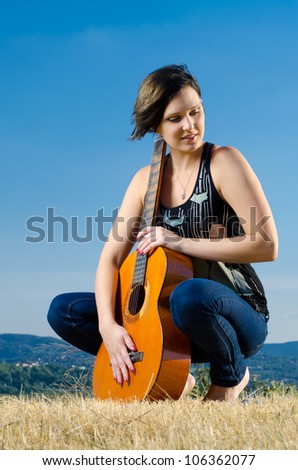 Beautiful attractive female guitarist posing with acoustic guitar on field against blue sky.